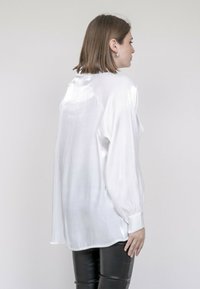 Blouse blanche brillante à manches longues et à coupe décontractée ; présente une texture lisse et un minimum de détails, associée à un pantalon noir ajusté.