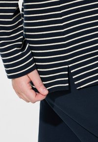 Marinblus med vita ränder, långärmad med rundad nederkant och sidoklipp. Tyget är mjukt och har en ribbad struktur. Hand som försiktigt håller i tyget.