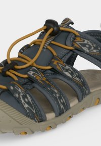 Κοντινή λήψη γκρι υπαίθριου σανδαλιού με υφασμάτινους λουριάδες με σχέδιο, ταμπά ελαστικά κορδόνια και ανθεκτική μπεζ σόλα με κίτρινες λεπτομέρειες.