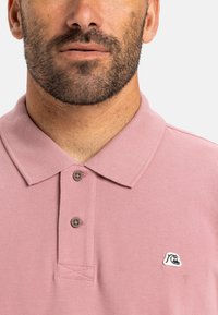 Růžová polo košile vyrobená z texturované látky s klasickým límcem, vybavená dvěma knoflíky a malým logem na levé straně.