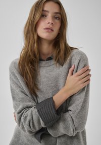 Grauer Strickpullover mit legerer Passform, versehen mit gerippten Bündchen und einem darunter hervorschauenden gestreiften Hemd. Weiche Textur, schlichtes Design.