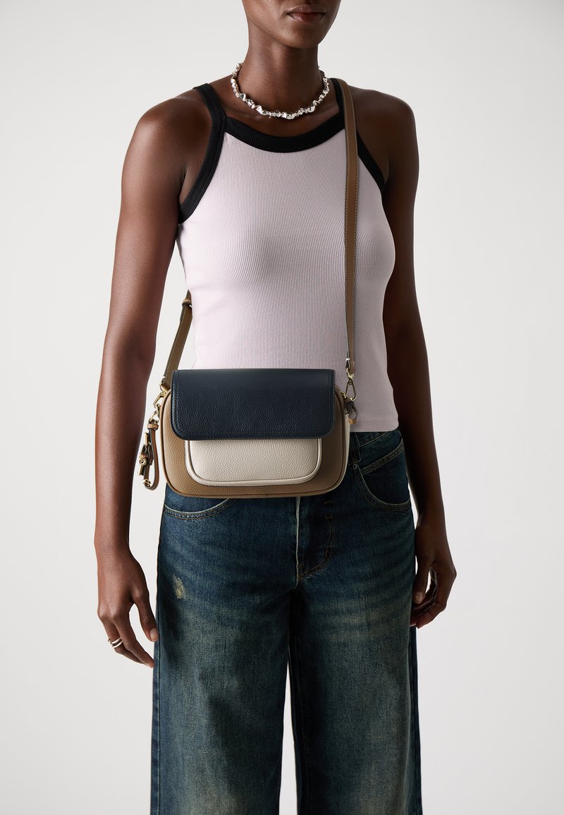 Leather crossbody bag with a navy top flap, cream bottom, and tan sides. Features gold hardware and a beige adjustable strap.