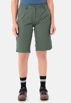 Personne portant un short kaki mi-long, des chaussettes noires à rayures blanches et des chaussures de randonnée grises, debout devant un fond uni.