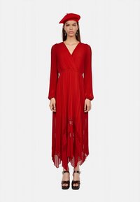 Rotes Faltenkleid mit langen Ärmeln und tiefem V-Ausschnitt. Der Saum ist asymmetrisch, und das Modell trägt ein passendes Barett sowie Plateau-Schuhe.