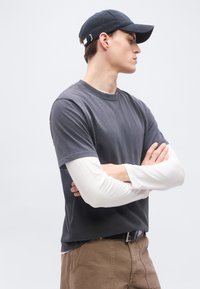 Νεαρός άνδρας που φοράει μαύρο καπέλο μπέιζμπολ, στρωμένα σκούρα γκρι και λευκά μακρυμάνικα πουκάμισα, καφέ παντελόνι, στέκεται με σταυρωμένα τα χέρια, κοιτάζοντας προς τα δεξιά.