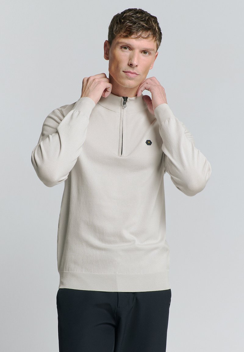 Beige zip-neck trui met lange mouwen, geribbelde manchetten en zoom, voorzien van een klein hexagonaal logo op de borst, gemaakt van zacht breimateriaal.
