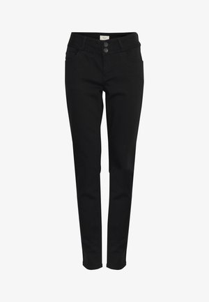 Femeie cu păr creț zâmbind, purtând un tricou alb, pantaloni negri și tocuri închise cu vârf ascuțit, stând în picioare pe un fundal simplu, deschis la culoare.