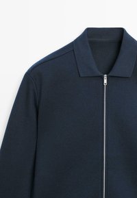 Mørkeblå lynlåstrøje med krave, der har en sølvfarvet lynlås og en glat tekstur. Skræddersyet pasform med minimale syninger.