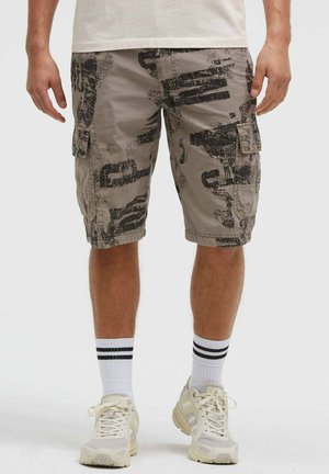 Mann trägt beige Cargo-Shorts mit schwarzem abstraktem Print, weiße Sportsocken mit schwarzen Streifen und beige Sneaker.