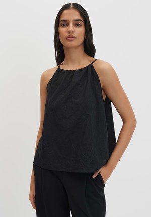 Vrouw met lang donker haar die een mouwloos zwart topje met subtiele bloemenborduur draagt en zwarte broek, staand met één hand in de zak.