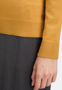 Mustár sárga pulóver bordás szegéllyel és mandzsettával, puha anyagból, sötét szürke ruhadarabbal párosítva.