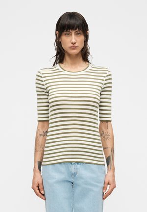 Person mit dunklem Haar, die ein olivfarbenes und weiß gestreiftes kurzärmeliges Hemd und hellblaue Jeans trägt, steht vor einem schlichten Hintergrund.