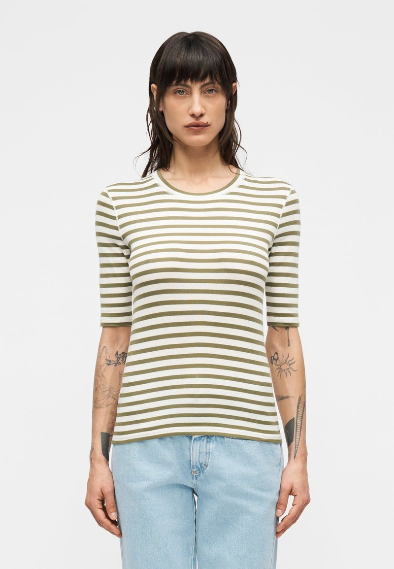 Άτομο με σκούρα μαλλιά που φορά μια κοντομάνικη ριγέ μπλούζα σε ελιά και λευκό και ανοιχτό μπλε τζιν, στέκεται μπροστά σε απλό φόντο.