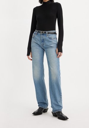 Person iført sort langærmet rullekrave, lyseblå jeans med højtaljet pas og sort bælte samt sorte spidse støvler, stående mod hvid baggrund.