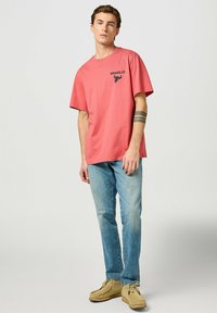 Camiseta rosa de manga corta con un logo negro de Wrangler, combinada con unos jeans de color azul claro y zapatos estilo mocasín en color beige.