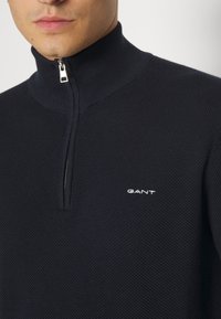 Navy zip-up pullover with a high collar, featuring a textured knit pattern and a small white GANT logo on the chest. Silver zipper hardware.