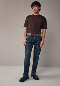 Brązowy bawełniany t-shirt o luźnym kroju, zestawiony z ciemnoniebieskimi dżinsami. Brązowy pasek i czarne buty. Prosty design, minimalne zdobienia.