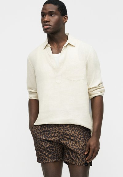 Homme portant une chemise crème à manches longues retroussées et un short marron à imprimé léopard, debout les mains dans les poches.