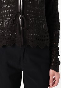 Brūns, atvērts adījums ar viļņotu apakšmalu un saišu aizdari, ar sarežģītiem rakstiem un teksturētām piedurknēm, savienots ar melniem biksēm.