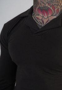 Tricou polo negru cu mânecă lungă și guler clasic. Material neted și elastic, care se potrivește strâns pe corp. Tatuaje notabile vizibile pe gât.