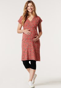 Vestido de maternidade com mangas curtas, decote em V, padrão floral em vermelho e preto, até ao joelho, combinado com leggings pretas e ténis brancos.