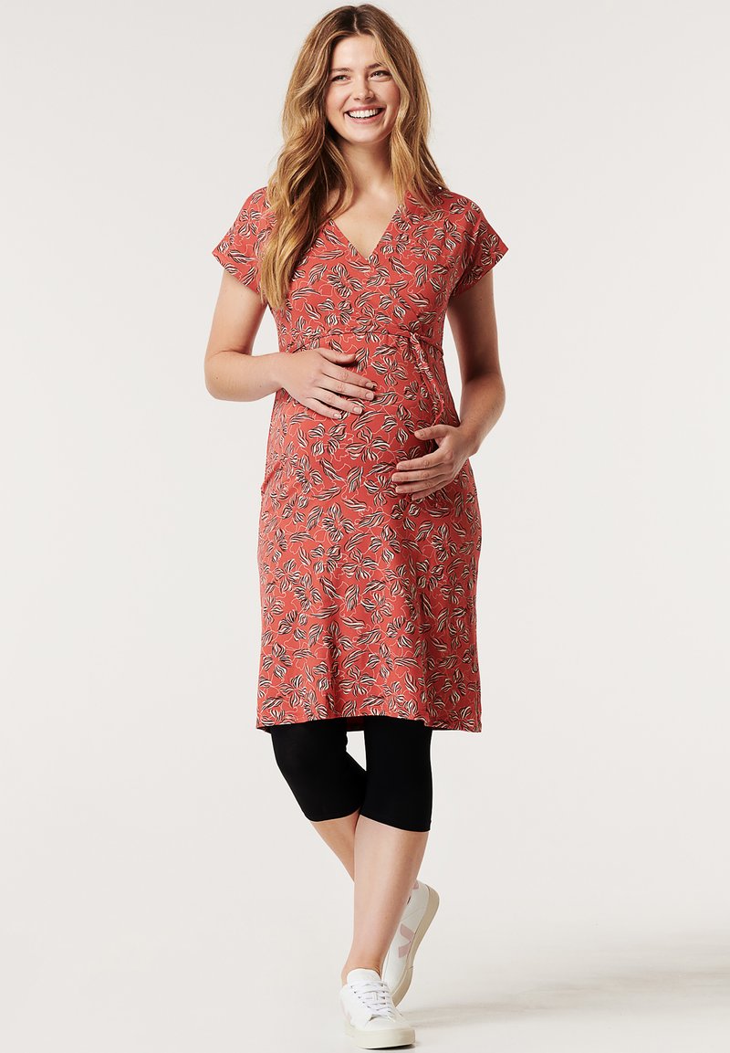 Vestido de maternidade com mangas curtas, decote em V, padrão floral em vermelho e preto, até ao joelho, combinado com leggings pretas e ténis brancos.