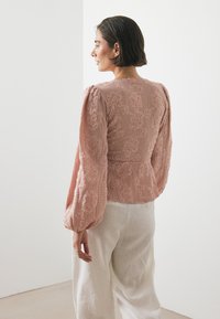 Blouse rosa tenue con texture, bordature floreali, maniche a sbuffo e vita realizzata su misura, abbinata a pantaloni in lino di colore chiaro.