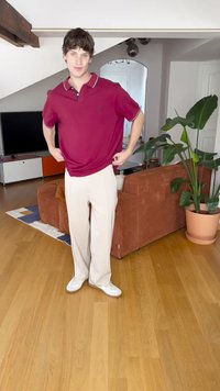 Rode polo shirt met kraag en contrasterende afwerking, gecombineerd met beige wijde broek. Witte sneakers, binnenomgeving met planten en meubels.