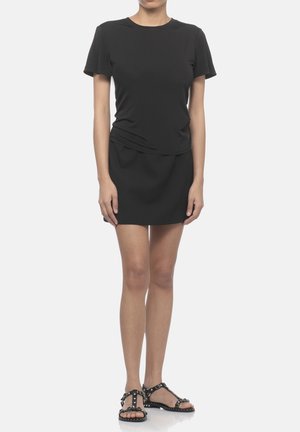 Kvinde iført en tætsiddende sort kortærmet kjole og sorte nitter flade sandaler, stående mod en ensfarvet hvid baggrund.