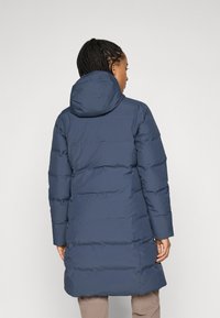 Persoon met gevlochten haar die een lange marineblauwe gewatteerde jas met capuchon draagt, staand met de rug naar de kijker.