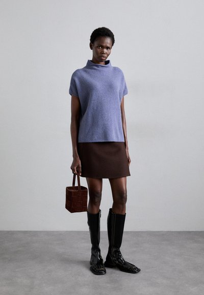 Top de lana de mangas cortas en color púrpura claro con cuello alto, combinado con una falda en línea A marrón y botas negras hasta la rodilla. Sostiene un bolso de mano tejido marrón.