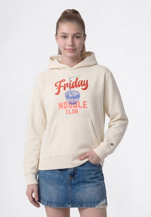 Млада жена с вързана назад коса носи кремаво худи с текст "Friday Noodle Club" и графика на синя купа с юфка, съчетано с дънкова пола.