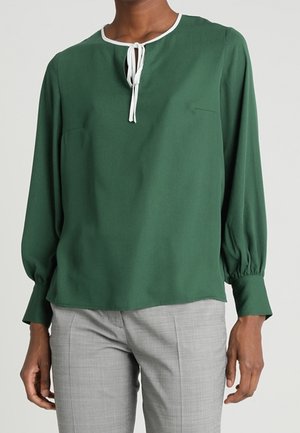 Henkilö, jolla on yllään tummanvihreä pitkähihainen paita valkoisella solmulla kaula-aukossa ja vaaleanharmaat housut, seisoo yksinkertaisen taustan edessä.