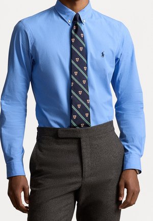 Muž v modré košili s knoflíky, s vzorovanou námořnicky modrou kravatou a tmavě šedými kalhotami s vysokým pasem na jednobarevném pozadí.