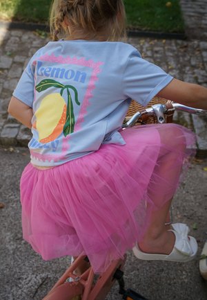 Ein Kind trägt ein blaues Hemd mit Zitronenprint und einen rosa Tüllrock und fährt draußen auf einem gepflasterten Weg ein pinkfarbenes Fahrrad.