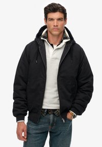 Zwarte hoodie met een voorrits en twee lagere zakken, gedragen over een lichtgrijs poloshirt met geruite accenten.