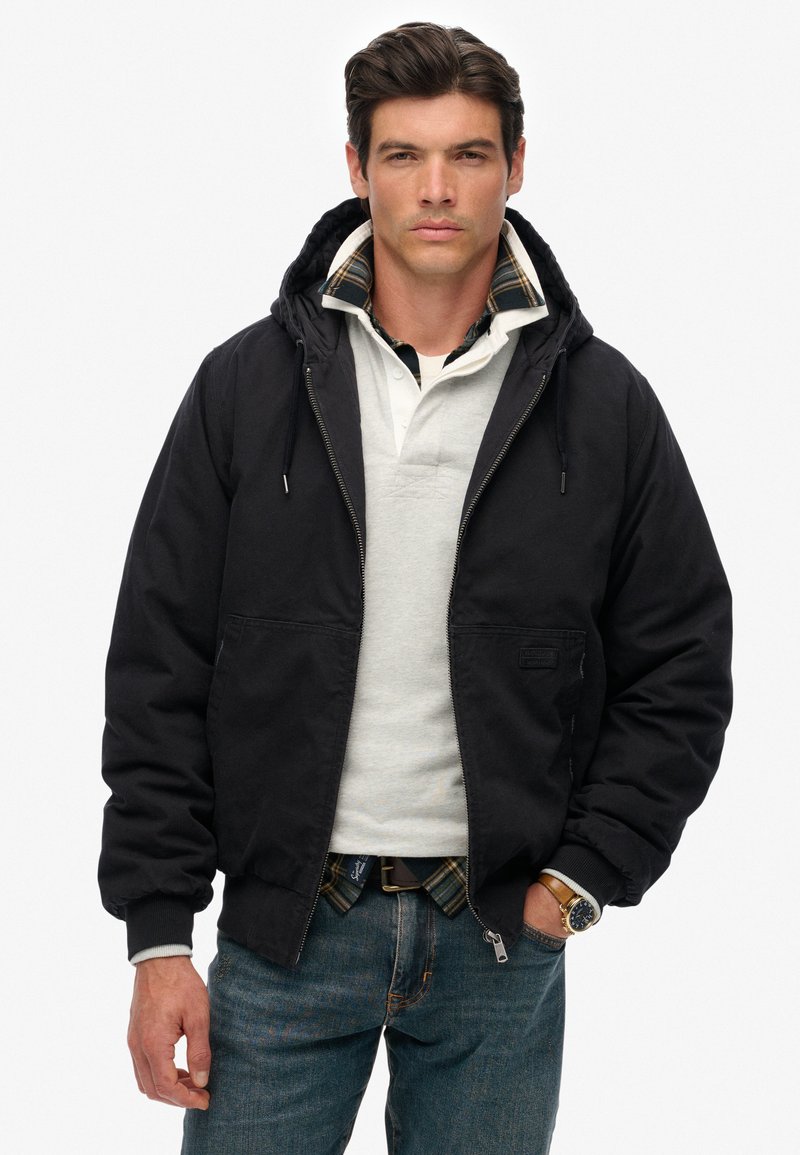 Zwarte hoodie met een voorrits en twee lagere zakken, gedragen over een lichtgrijs poloshirt met geruite accenten.