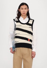 Sweter bez rękawów w kolorze kremowym i czarnym, z wzorem w zygzaki oraz haftem czerwonej truskawki. Połączony z beżowymi spodniami.