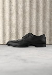 Chaussure habillée brogue en cuir noir avec perforations décoratives, lacets et talon bas, montrée de profil sur une surface claire texturée et un fond à rayures verticales.