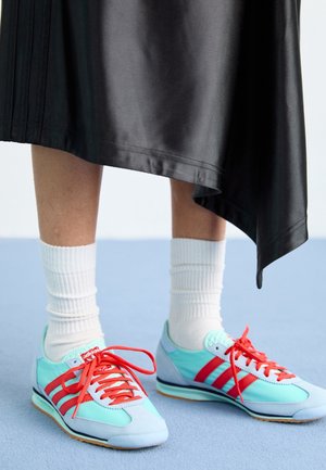 Benedenbenen met witte geribbelde sokken en turquoise en rode Adidas sneakers met rode veters, staand op een lichtblauwe vloer onder een zwarte asymmetrische rok.