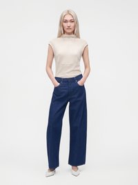 Жена с дълга руса коса, носеща кремав безръкав топ, тъмносини широки дънки, бели заострени обувки, стояща с ръце в джобовете.