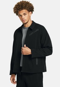 Chaqueta negra con cremallera y corte relajado, que presenta un pequeño bolsillo con cremallera en el pecho y un tejido texturizado. Se lleva sobre una camisa gris.