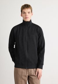 Giacca nera con zip caratterizzata da un motivo geometrico testurizzato, colletto alto e polsini e orlo a costine. Abbinata a pantaloni beige a vestibilità rilassata.