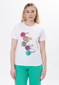 Camiseta blanca con un gráfico de cinco anillos con piedras coloridas: rosa, púrpura, turquesa, negro y acentos dorados, en un modelo.