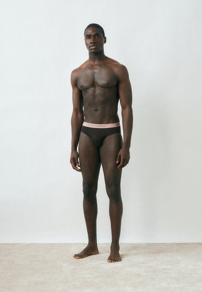 Μυώδης άνδρας με σκούρο δέρμα, όρθιος ξυπόλητος πάνω σε χαλί μπροστά σε απλό ανοιχτόχρωμο τοίχο, φορώντας σκούρα εσώρουχα με ανοιχτό ροζ ζώνη μέσης.