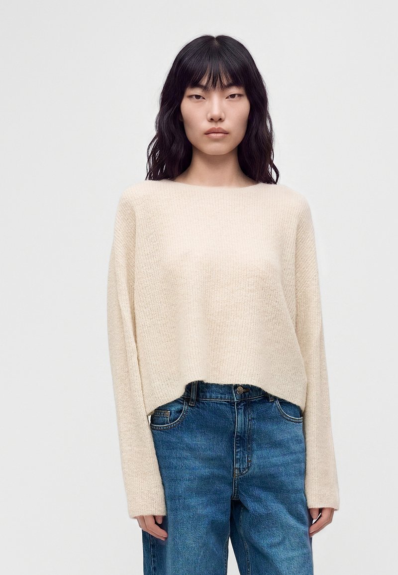 Femme aux longs cheveux noirs portant un pull beige ample et un jean bleu taille haute, debout devant un fond clair uni.