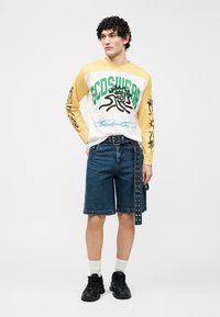 Άνδρας που φοράει μακρυμάνικο γραφιστικό πουκάμισο σε κίτρινο και λευκό, μπλε τζιν σορτς με μαύρη ζώνη, λευκές κάλτσες και μαύρα αθλητικά παπούτσια, στέκεται μπροστά σε λευκό φόντο.