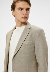 Beige texturerad blazer med slag och en enda bröstficka, bärs över en ljusfärgad rundhalsad tröja. Modellen står med sidan lite vänd.