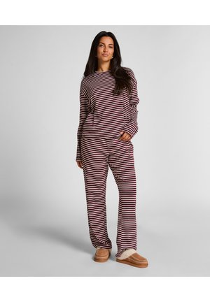 Femme se tenant debout, portant un ensemble assorti de haut et de pantalon rayés bordeaux et blancs à manches longues, avec des pantoufles en fourrure beige sur un fond gris.