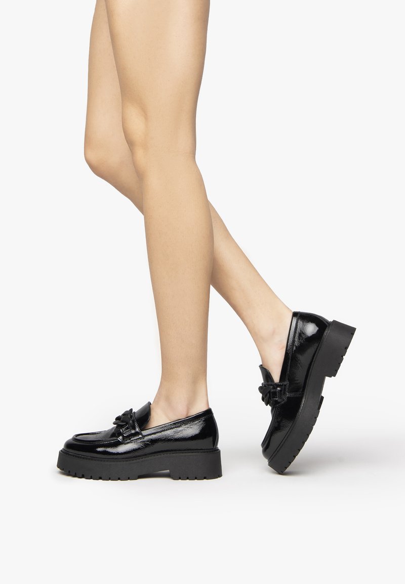 Mocassins noirs en cuir verni avec une semelle épaisse, détail de nœud et texture lisse, mettant en avant un design décolleté et des lignes épurées.